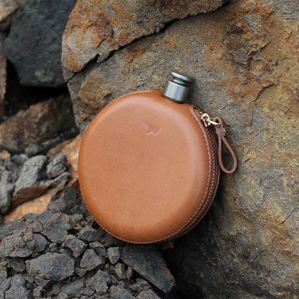 Round titanium flask with brown leather cover on rocks