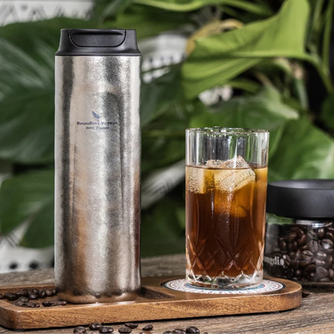 Titanium French press mug beside a glass of coffee on outdoor table, ideal for travel or campsite brewing