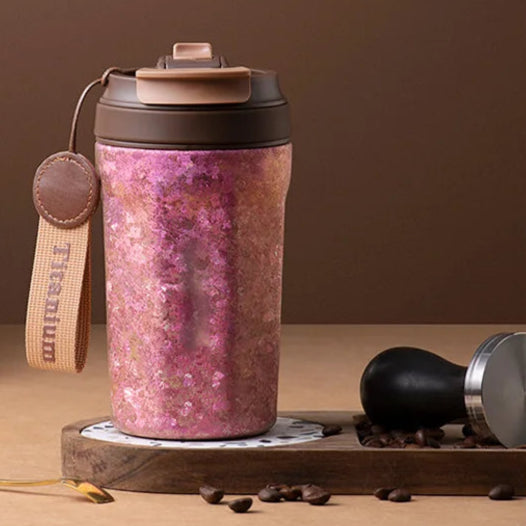Pink titanium thermos cup with leather strap and straw lid placed on wooden desk beside coffee accessories