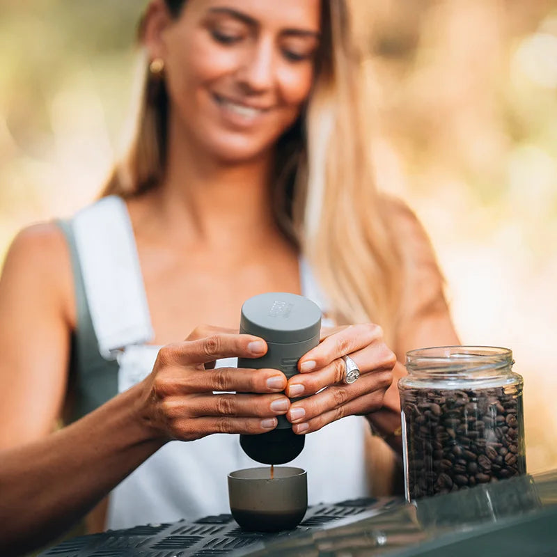 Woman preparing espresso outdoors with portable Minipresso coffee maker on campsite table